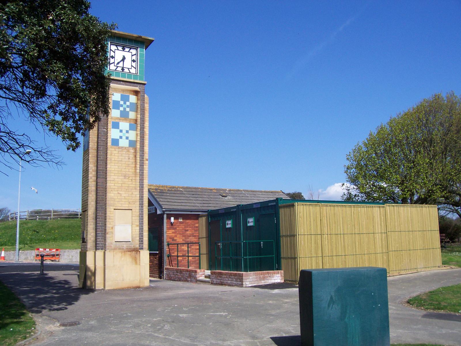 Green modular public toilet units beside a seafront clock tower with timber screening